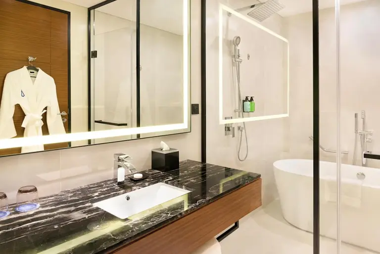 Asiana Grand Hotel Dubai Badezimmer mit Marmorwaschtisch, beleuchtetem Spiegel, Glasdusche und Badewanne