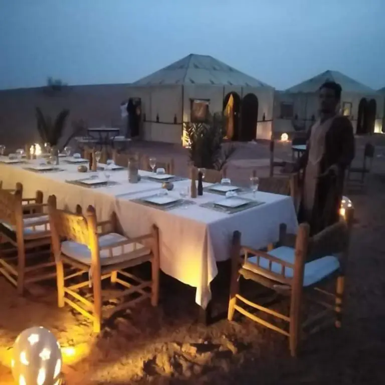 Abendliches Dinner-Setup im Berber Soul luxury Camp in Merzouga bei Dämmerung mit beleuchteten Zelten und Kerzenlicht