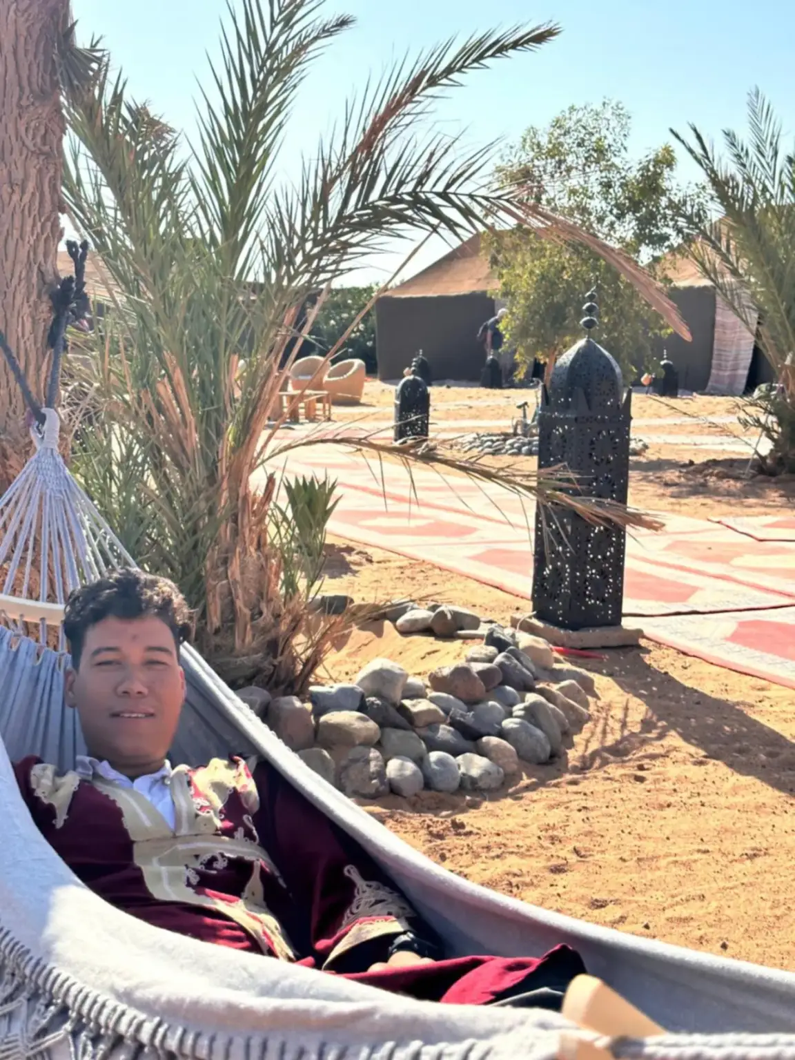 Eco Merzouga luxury camp in Merzouga with a guest reclining in a hammock near palm trees and desert paths