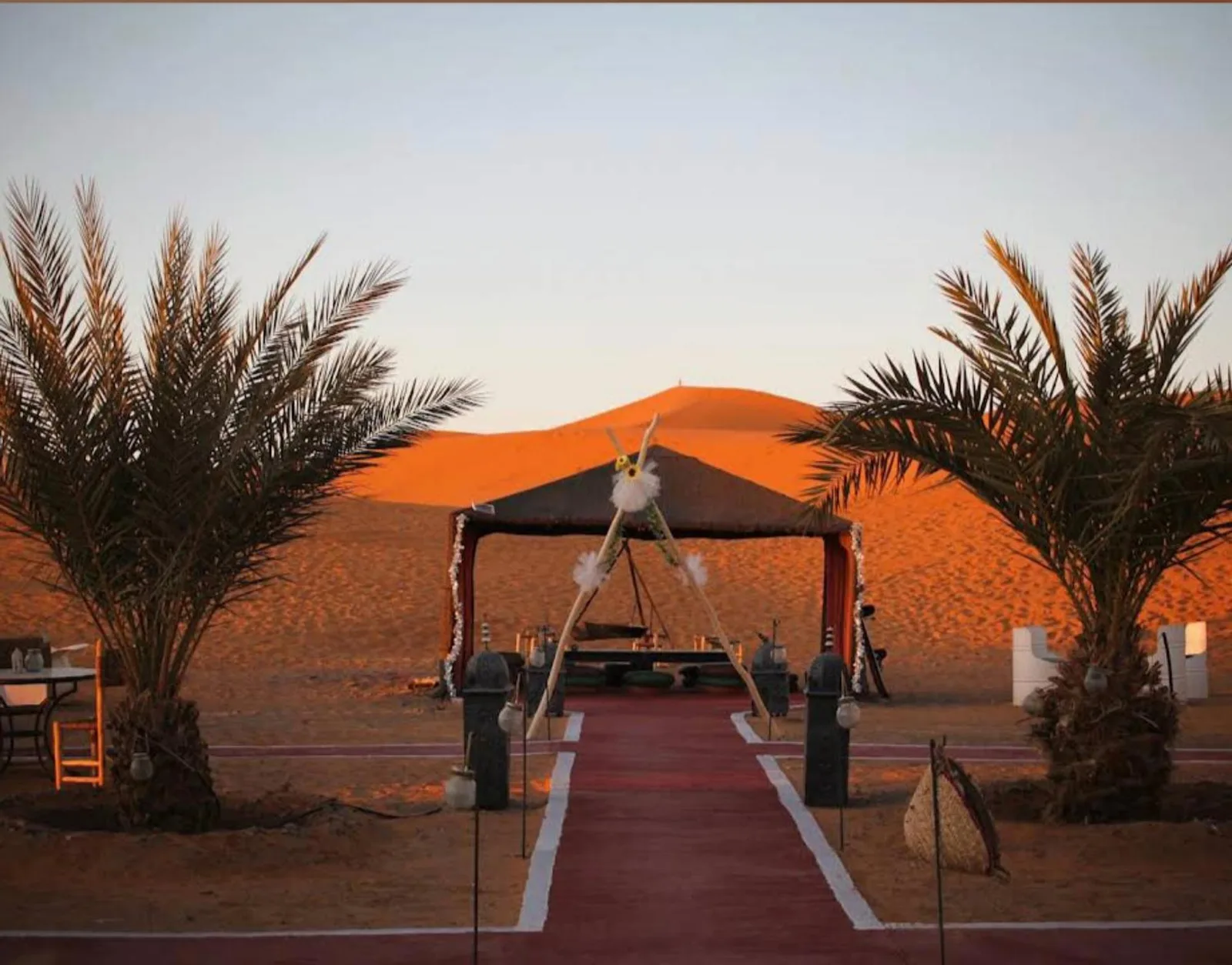 Entrada exterior de Erg Dream Luxury Camp en Merzouga, con palmeras y dunas de arena al atardecer.