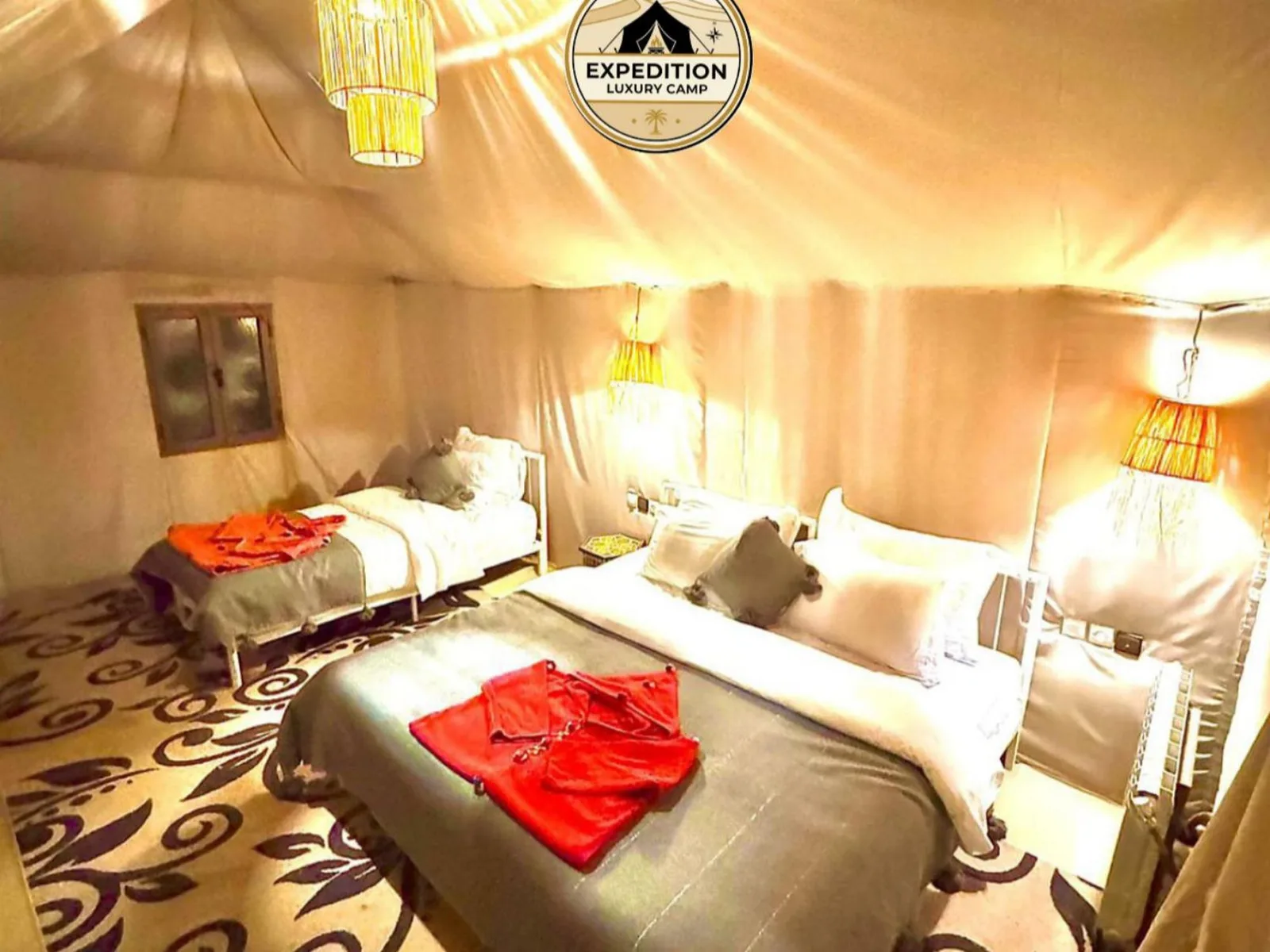 Habitación familiar con tienda de Expedition Luxury Camp en Merzouga con varias camas, iluminación cálida y alfombras estampadas en el suelo