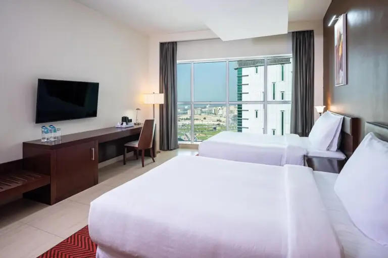 Twin guest room at Four Points by Sheraton Sharjah with two beds, desk, and large city-view window.