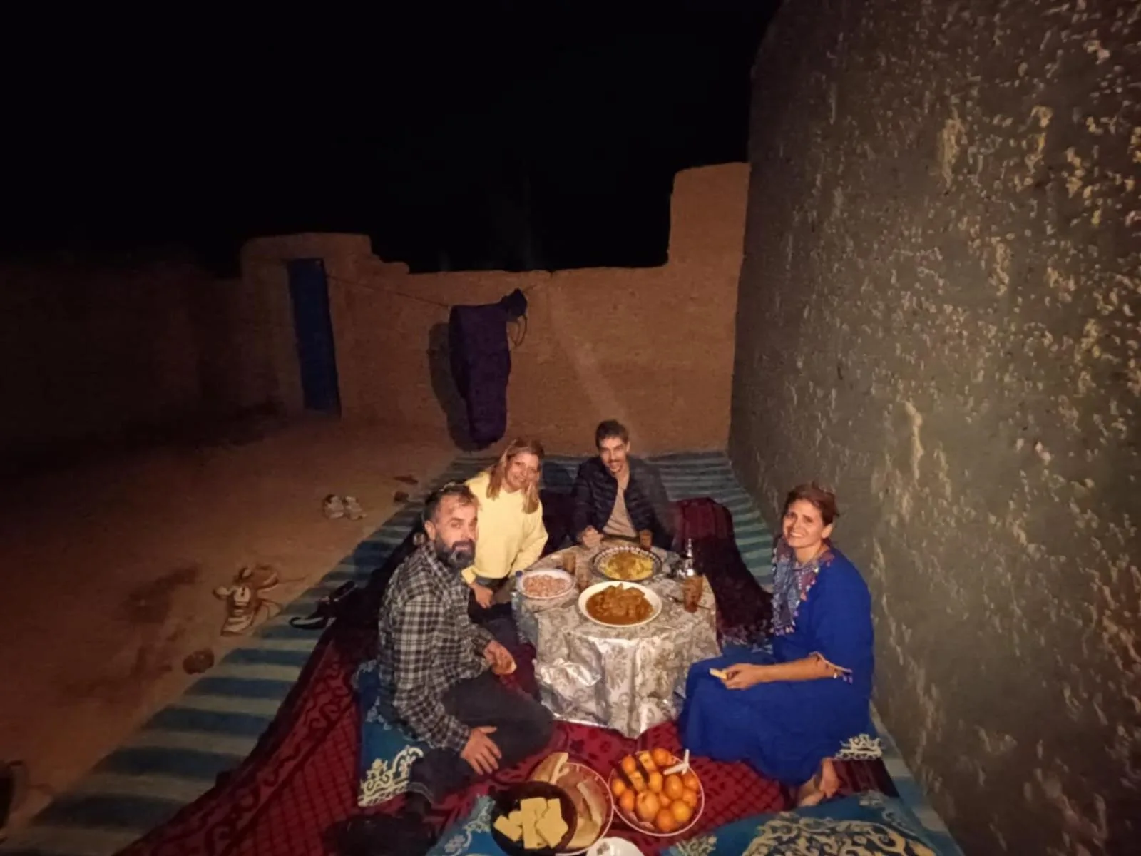 Abendessen im Freien bei Nacht im Guest House Nomad Life in Merzouga mit Gästen auf Teppichen neben einer Wand.