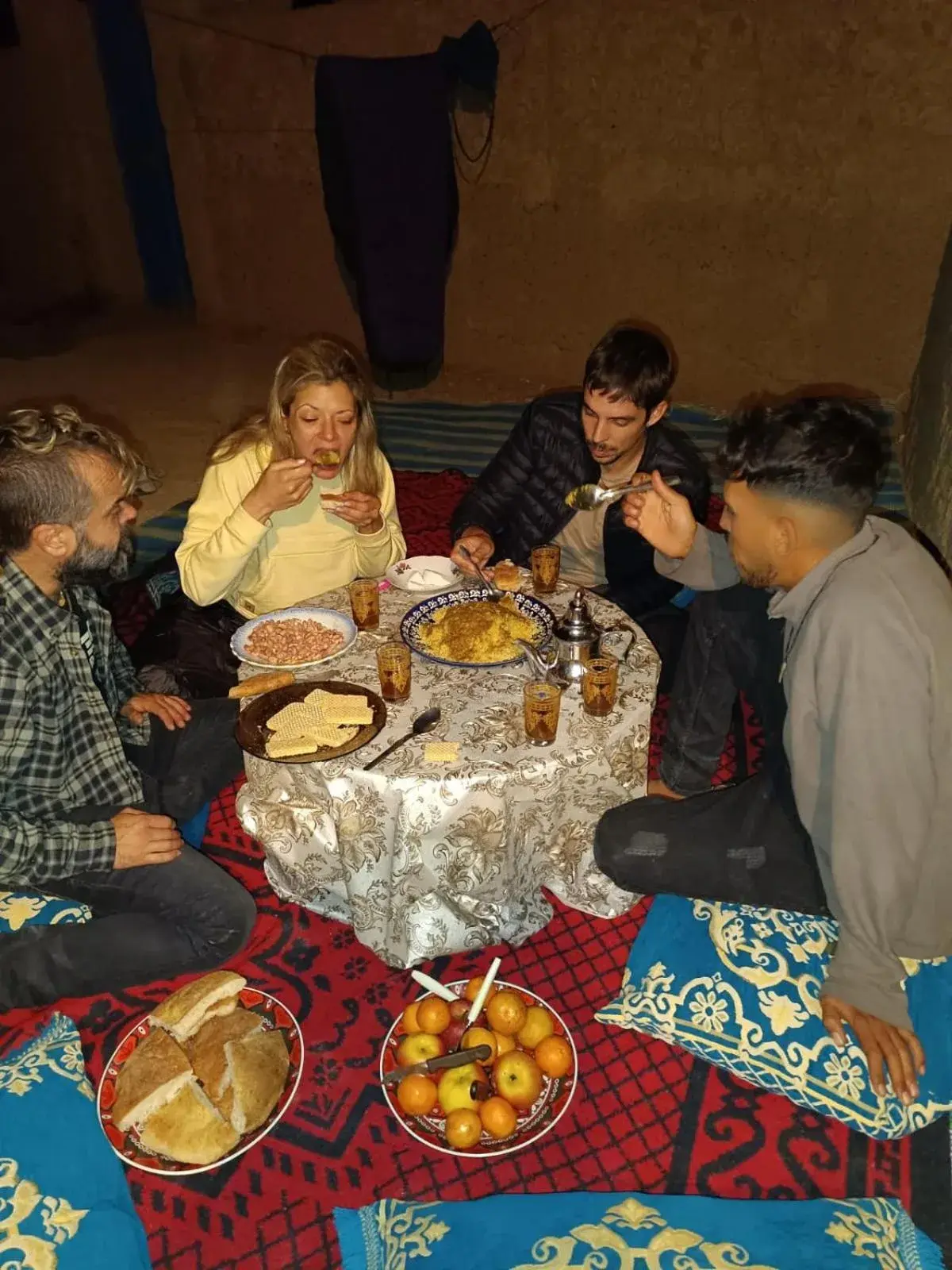 Gäste beim gemeinsamen traditionellen Abendessen im Guest House Nomad Life in Merzouga an einem niedrigen Tisch auf gemusterten Teppichen.