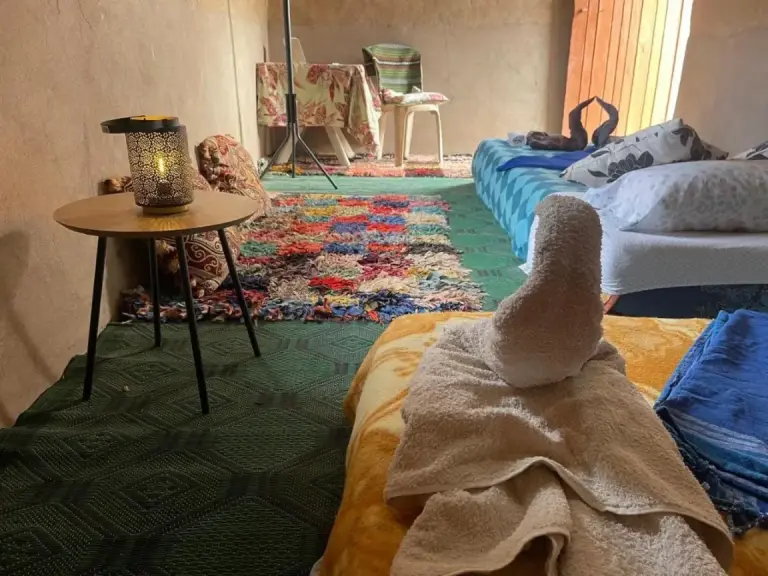 Gemütliches Gemeinschaftsgästezimmer im Guest House Nomad Life in Merzouga mit Betten, Teppichen und einer kleinen Tischlampe.