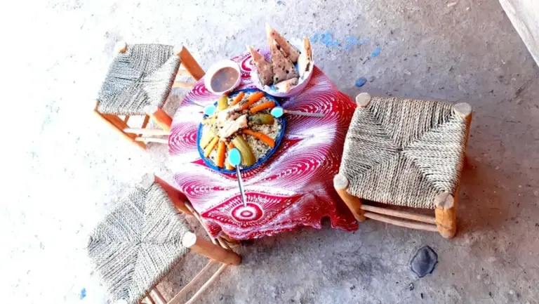 Outdoor low table setup at Guests House in Merzouga with woven stools and shared food in a rustic setting.