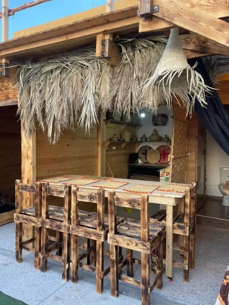 Outdoor dining area at Heaven Surf Camp in Agadir with wooden stools, thatched roof, and serving counter