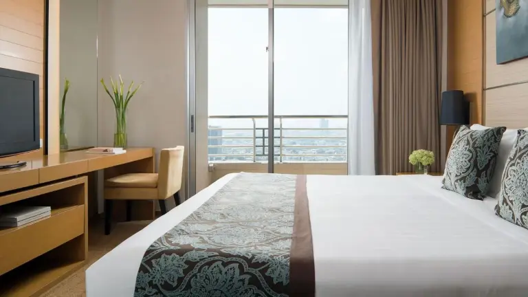 Bedroom interior at JC Kevin Sathorn Bangkok Hotel with a bed, desk, and city view through the window.