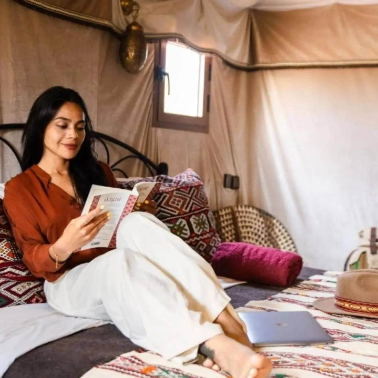 Gast liest in einem Zelt im Lana Camp Merzouga in Merzouga mit gemusterten Kissen und Laptop in der Nähe.