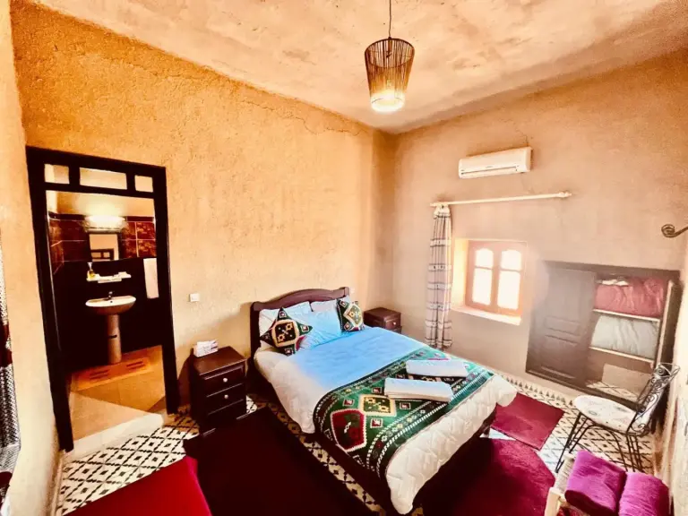 Una habitación espaciosa en el hotel Les Roses Des Dunes de Merzouga con una cama doble, ventana, acceso al baño y baldosas tradicionales en el suelo.
