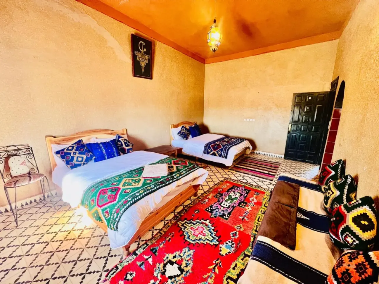 Una habitación triple en el hotel Les Roses Des Dunes de Merzouga con dos camas, alfombras coloridas y cojines de estilo bereber.