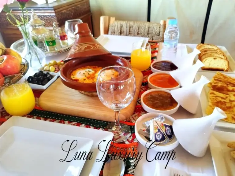 Table de petit-déjeuner à Luna Luxury Camp Merzouga à Merzouga avec tajine, fruits, jus, pain et service de thé.