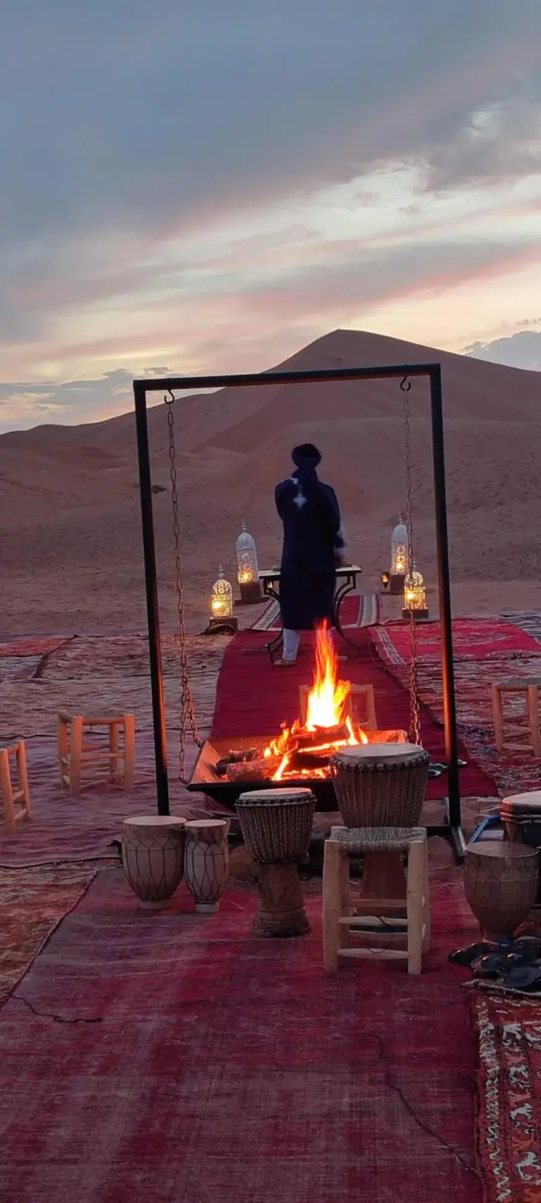 Espace feu de camp au coucher du soleil à Luna Luxury Camp Merzouga à Merzouga avec tapis, tambours, lanternes et assises dans le désert.