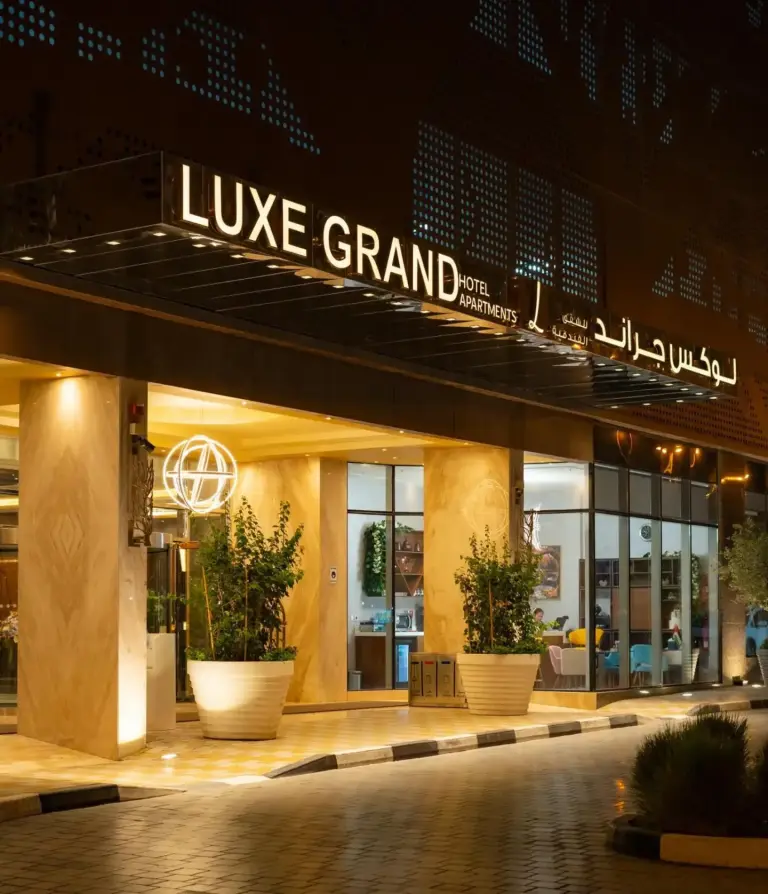 Nachtansicht des Eingangs zum Luxe Grand Hotel Apartments in Schardscha mit beleuchtetem Schild und gläserner Lobbyfront.