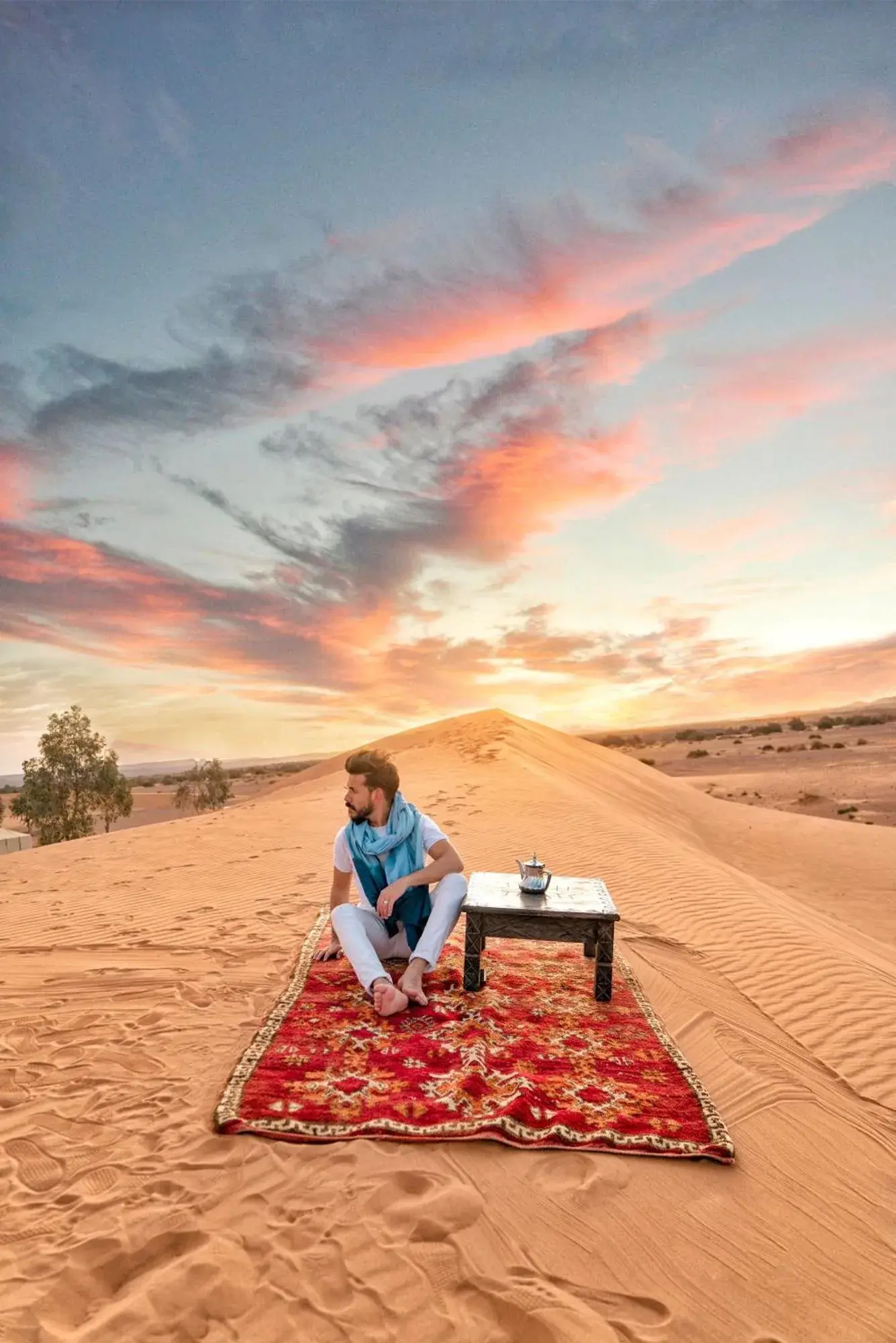 Scène de coucher de soleil dans le désert au Luxury Merzouga Stars Camp à Merzouga, avec un client assis sur un tapis au sommet des dunes.