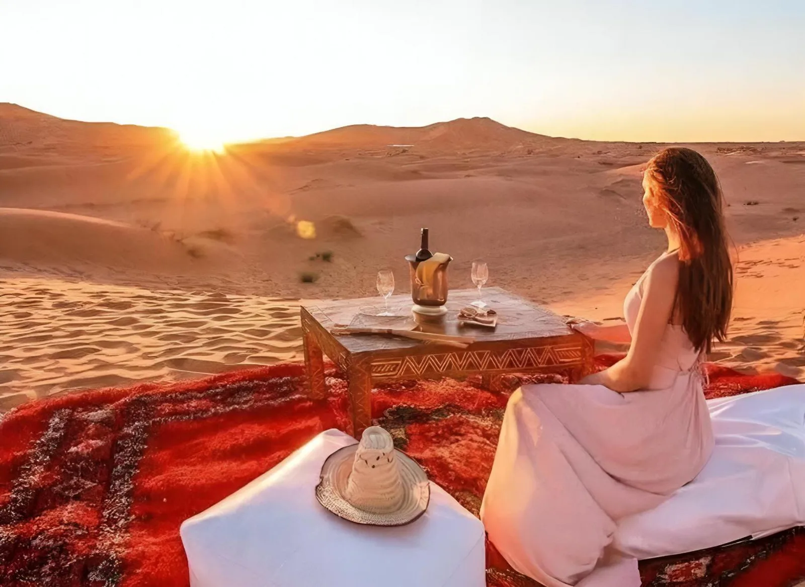 Installation de dîner au coucher du soleil au Luxury Merzouga Stars Camp à Merzouga avec un client assis sur un tapis dans le désert.