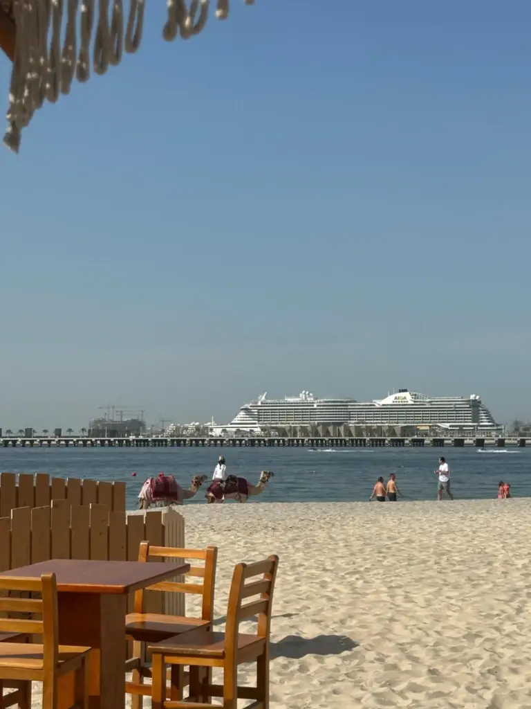 Vista de la playa desde Magic JBR & Dubai Marina en Dubai con una orilla de arena, asientos de un café junto al mar y un crucero mar adentro.