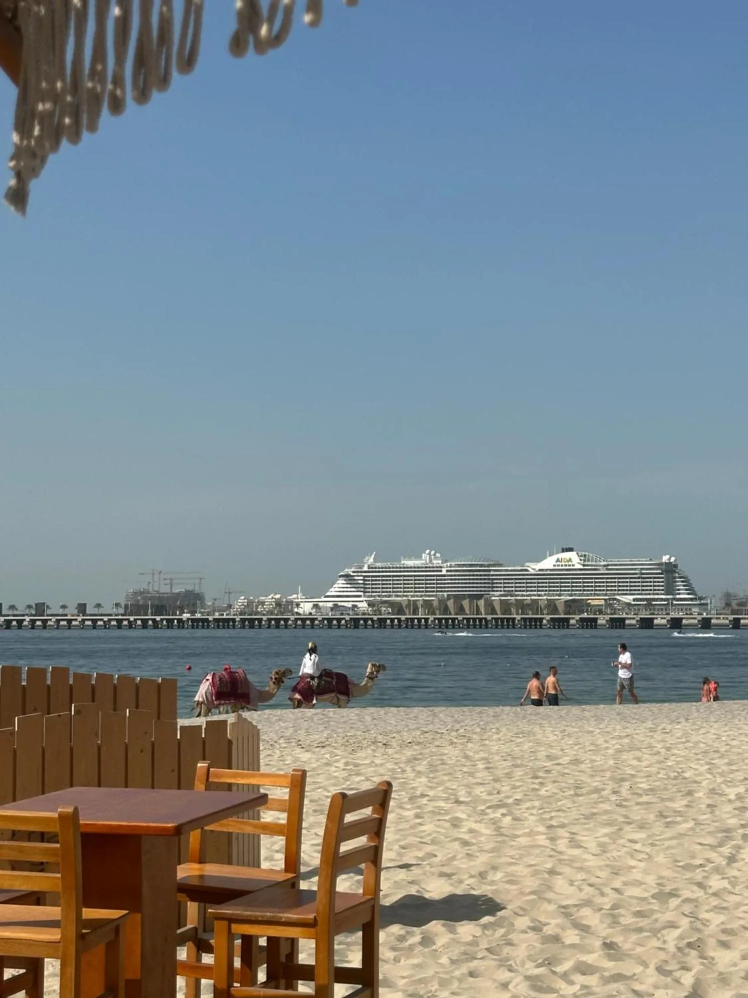 Vista de la playa desde Magic JBR & Dubai Marina en Dubai con una orilla de arena, asientos de un café junto al mar y un crucero mar adentro.
