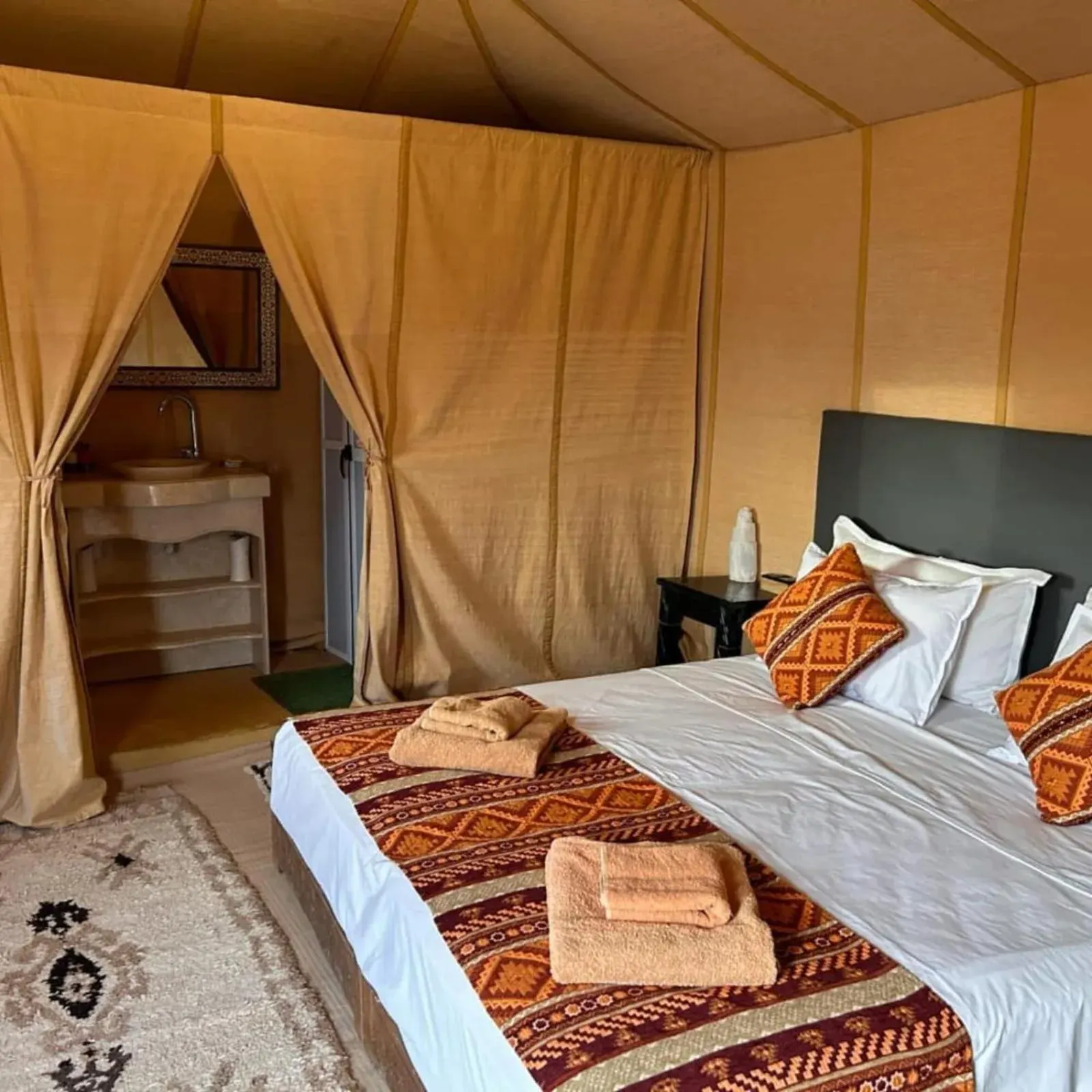 Elegantes Doppelzeltzimmer im Magnifique Luxury Camp in Merzouga mit großem Bett, Teppichen und Eingang zum Badezimmer.
