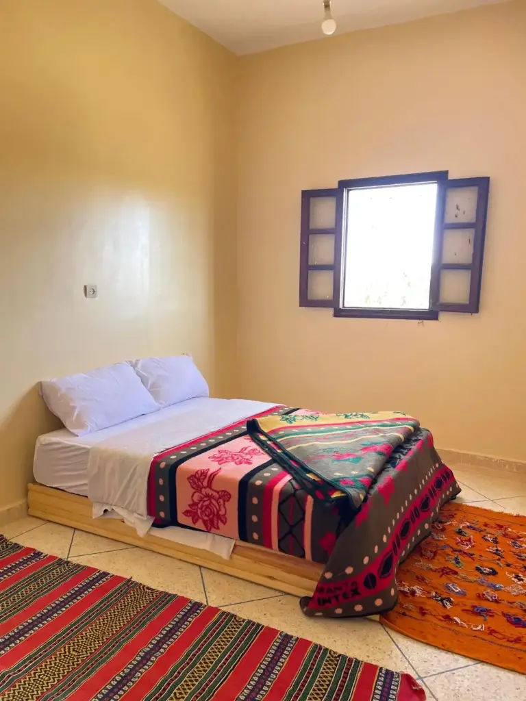 Double room at Maison La Baraka Merzouga in Merzouga with a bed, colorful blanket, patterned rug, and window.