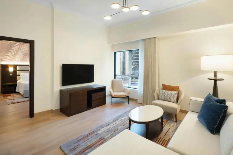 Wohnzimmer einer Suite im Marriott Hotel Production City, Dubai mit Sofa, TV, Stühlen und Fenster zur Stadtseite