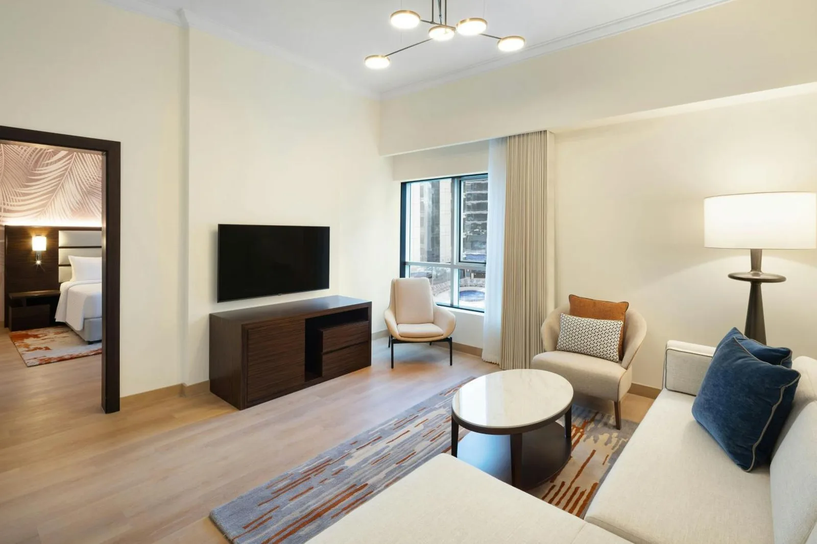 Wohnzimmer einer Suite im Marriott Hotel Production City, Dubai mit Sofa, TV, Stühlen und Fenster zur Stadtseite
