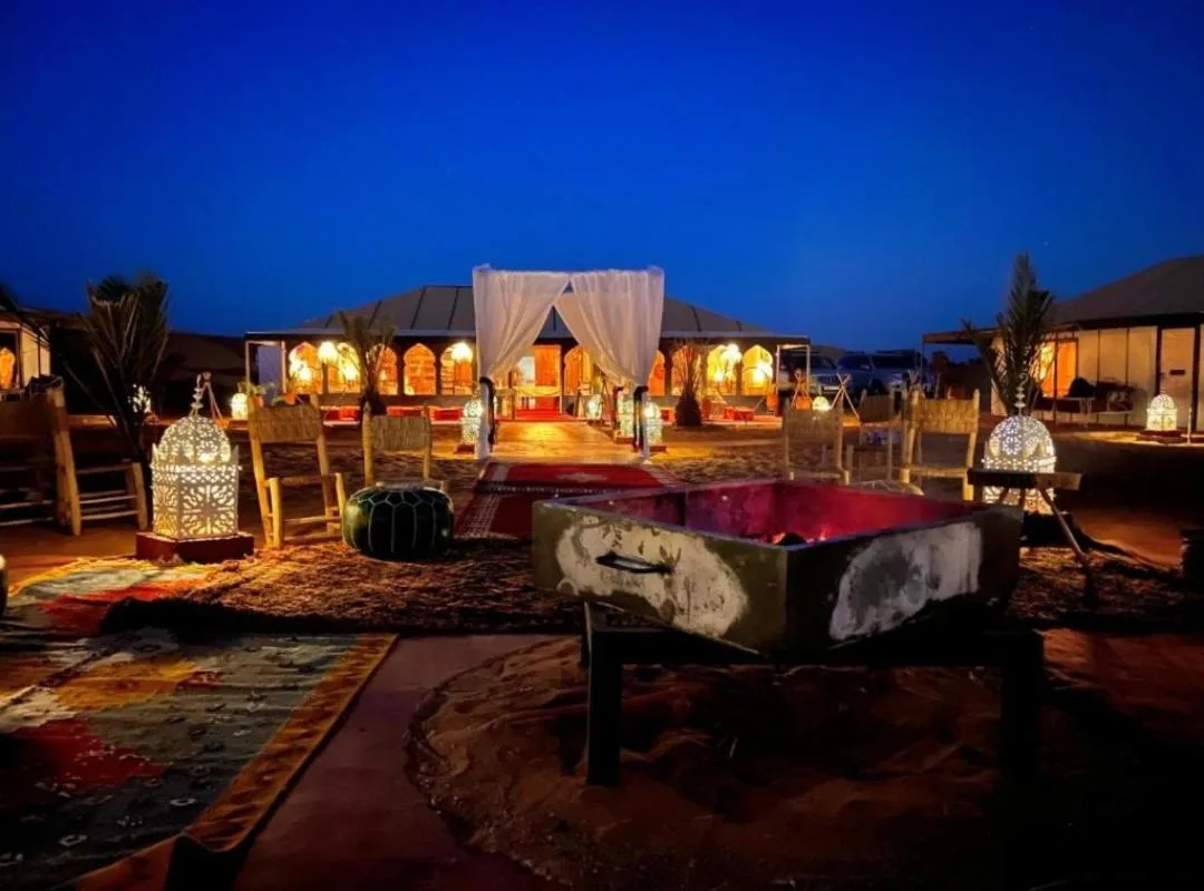 Night exterior of Merzouga Aicha Luxury Camp in Merzouga with illuminated entrance and desert seating.