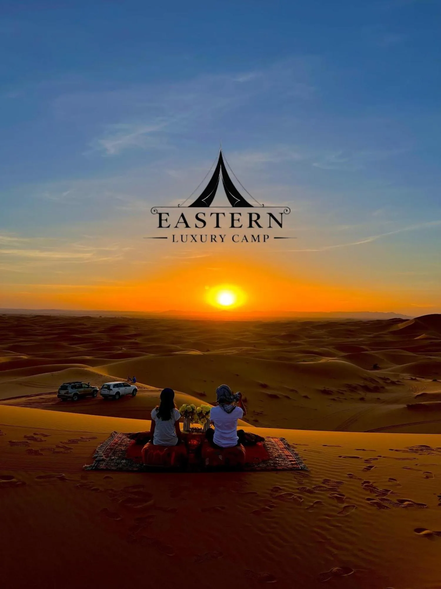 Atardecer en el desierto en Merzouga Eastern Camp Luxury en Merzouga con huéspedes sentados sobre una alfombra con vistas a las dunas.