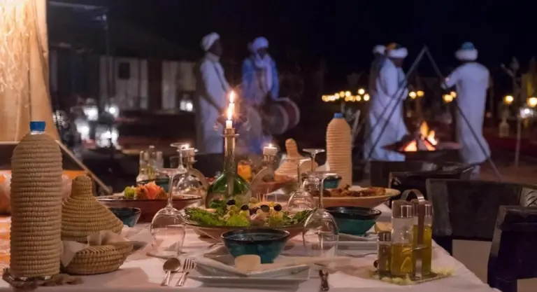 Abendliches Dinner-Setup im Merzouga Sara Luxury Camp in Merzouga mit Tischservice und Lagerfeuer im Hintergrund