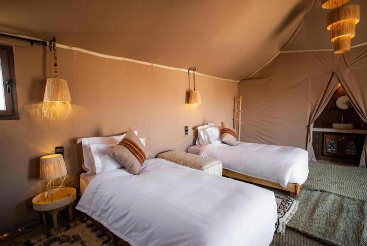 Gästezelt mit zwei Betten im Merzouga Sara Luxury Camp in Merzouga mit warmem Licht und traditioneller Wüsten-Dekoration