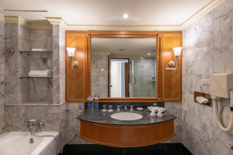 Bathroom vanity at Montien Riverside Hotel Bangkok with marble countertop, large mirror, bathtub, and toiletries.