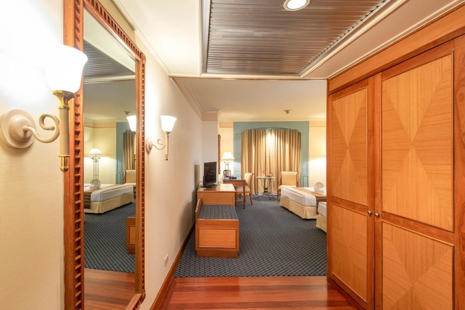 Suite interior at Montien Riverside Hotel Bangkok with wardrobe, mirror, seating area, and twin beds in the distance.