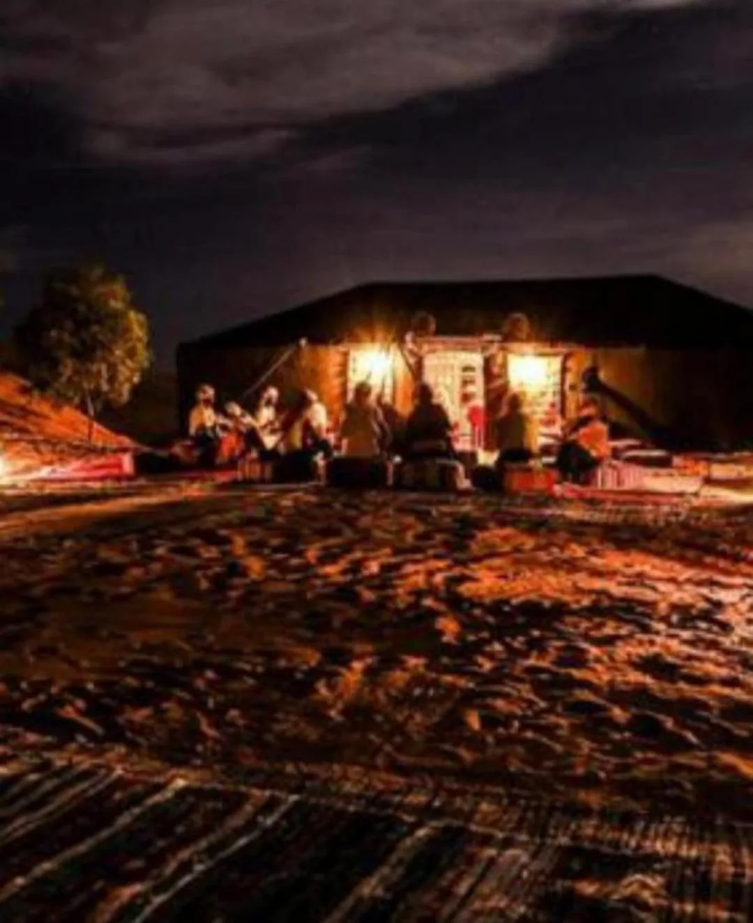 Vue extérieure de Overnight Desert Camp à Merzouga la nuit avec des clients à l’extérieur du camp illuminé.