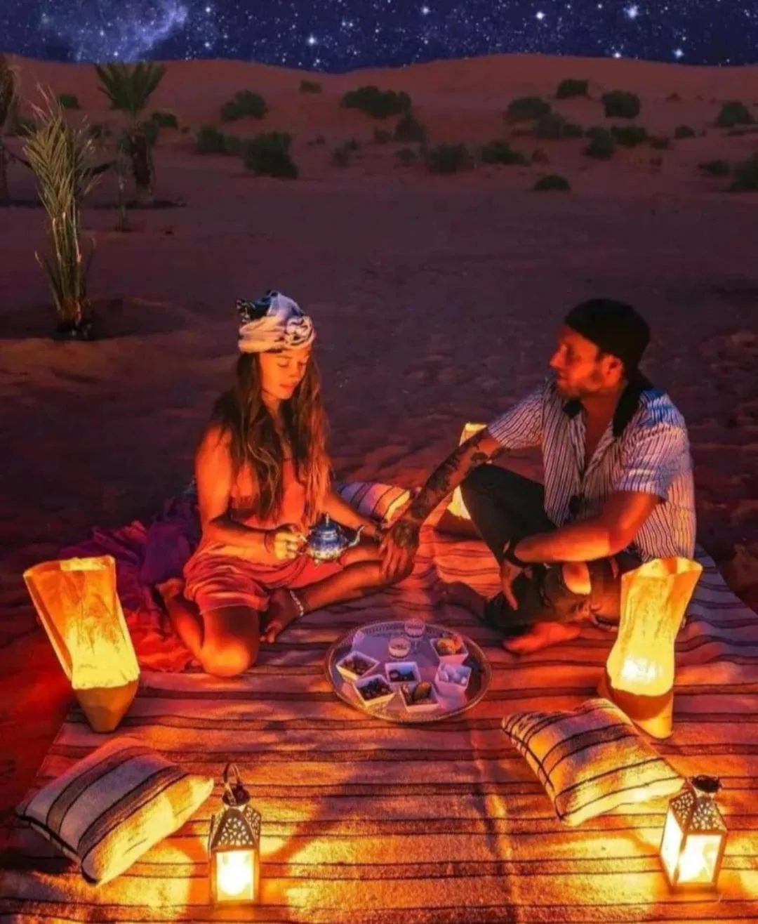 Une installation romantique dans le désert à Overnight Desert Camp à Merzouga avec des lanternes, des coussins et un couple assis sur un tapis.