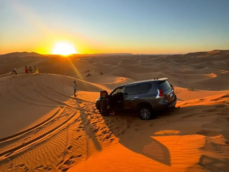 Un véhicule 4x4 garé sur les dunes au coucher du soleil près de Premium Luxury Camp à Merzouga