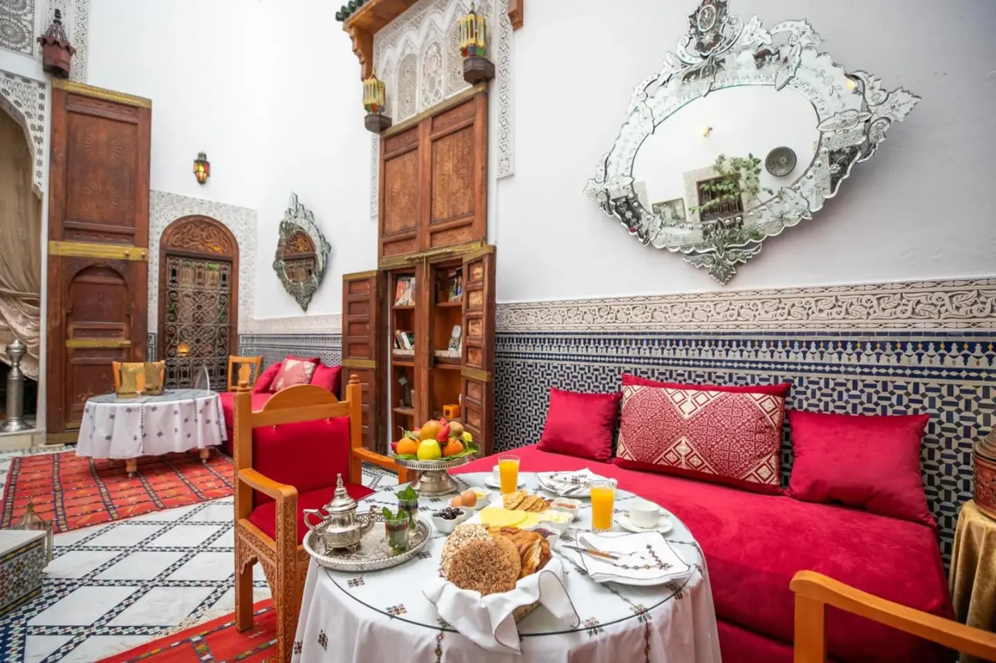 Courtyard lounge at Riad Arabella in Fes with red sofa, breakfast table, carved mirror, and mosaic decor.