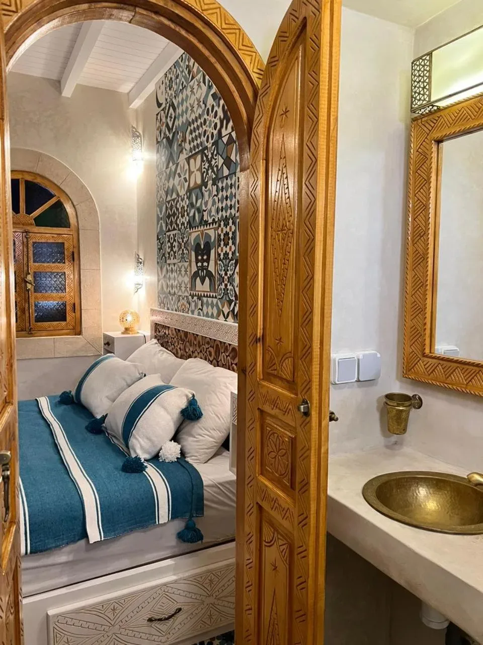 Entrada al baño en Riad dar el hania en Essaouira con puerta de madera tallada, fregadero de latón y dormitorio al fondo