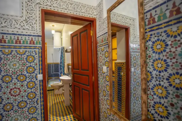 Baño en Riad Fes iline Medina en Fes con coloridos azulejos de mosaico, un inodoro, lavabo, espejo y puerta de madera