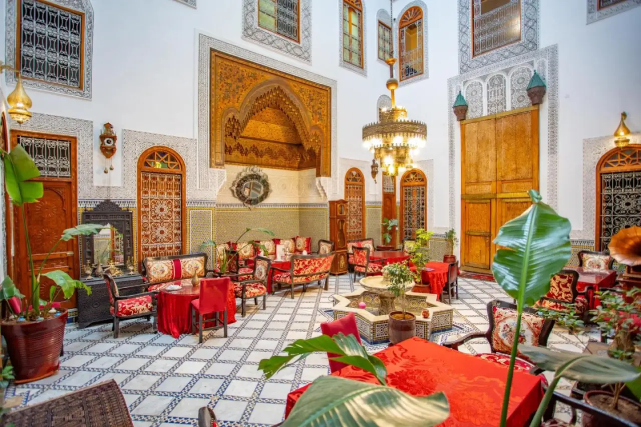 Salón del patio en Riad Fes iline Medina en Fes con asientos marroquíes ornamentados, mosaicos y una lámpara de araña central