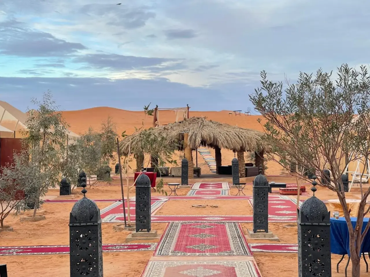Roses desert camp in Merzouga courtyard with patterned carpets, shaded seating, and desert dunes in the background