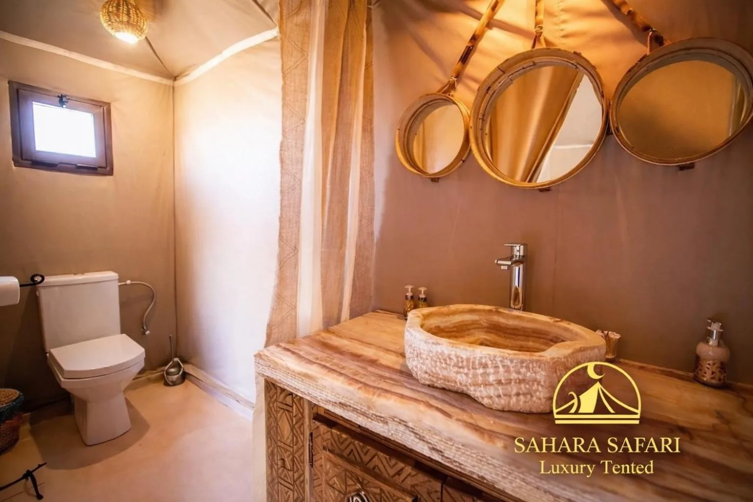 Badezimmer im Sahara Safari Luxury Camp & Activities in Merzouga mit Steinwaschbecken, Toilette und runden Spiegeln in einem warmen Innenraum.