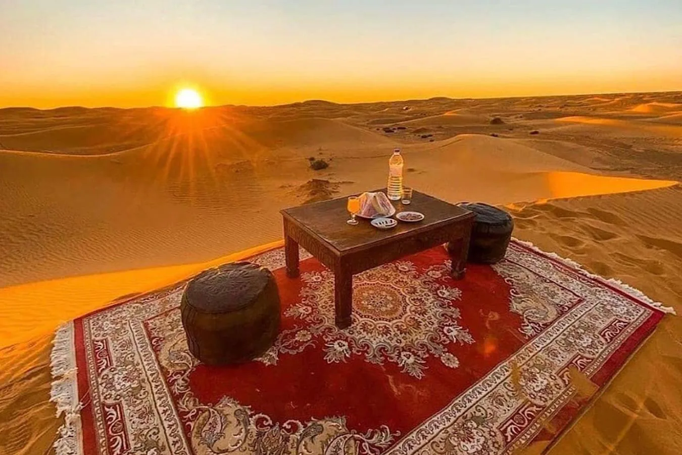 Desert sunrise dining setup at Soul Dunes Luxury Camp in Merzouga with a low table, rugs, and cushions on the sand dunes
