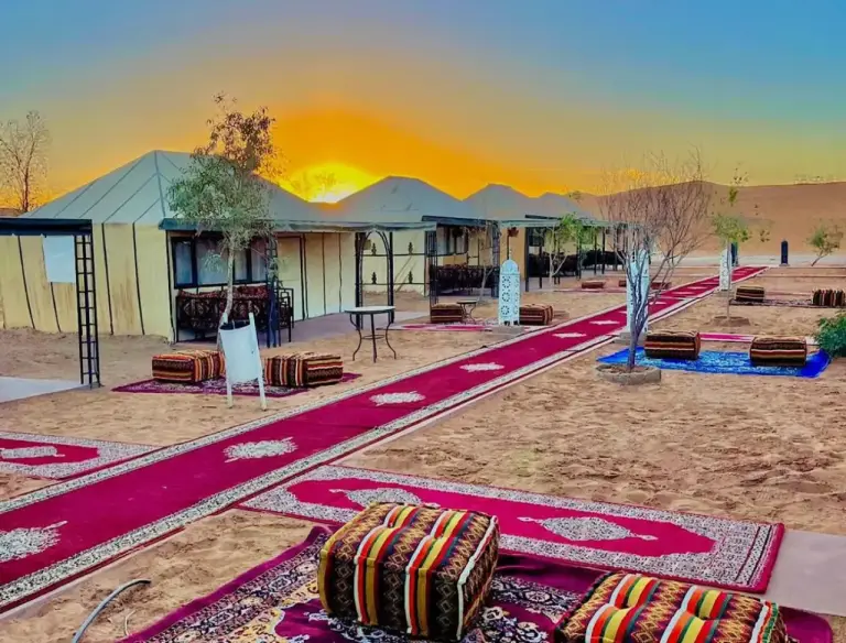 Sunshine luxury tent in Merzouga desert pathway with colorful carpets, lounge seating, and sunrise light across the camp.