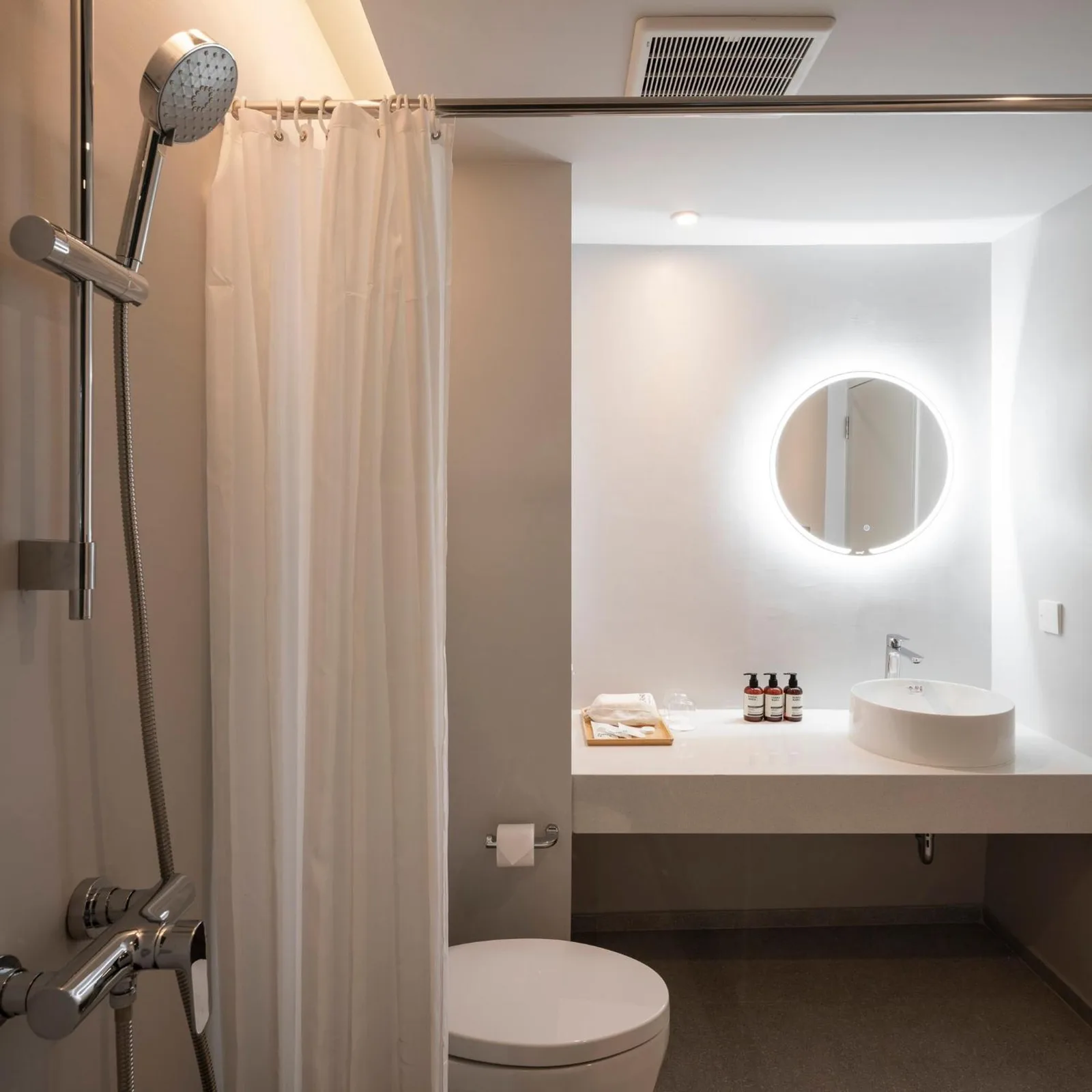 Modernes Badezimmer im Hotel Terra Nara Pattaya mit Dusche, Toilette, Waschtisch und rundem beleuchtetem Spiegel.