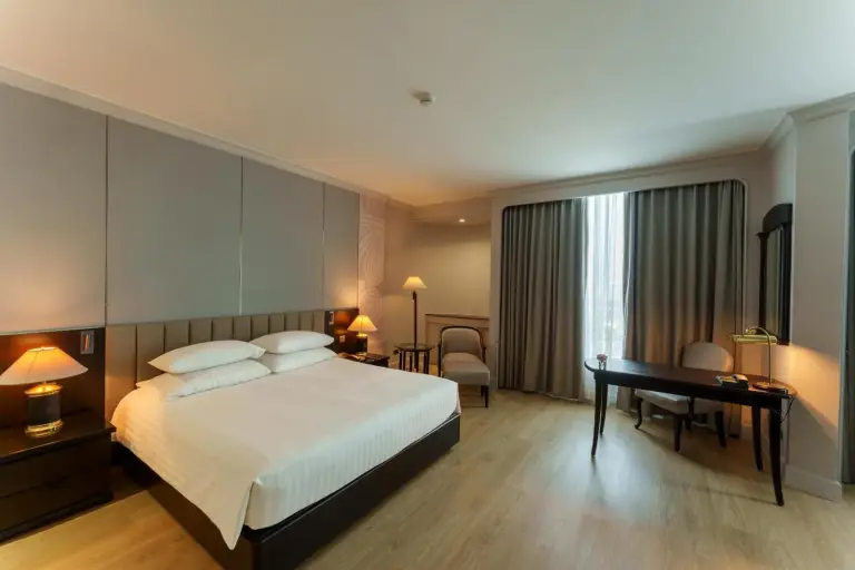 Deluxe-Schlafzimmer im The Heritage Grand Khon Kaen Hotel and Convention in Khon Kaen mit Kingsize-Bett, Schreibtisch, Vorhängen und warmer Beleuchtung.