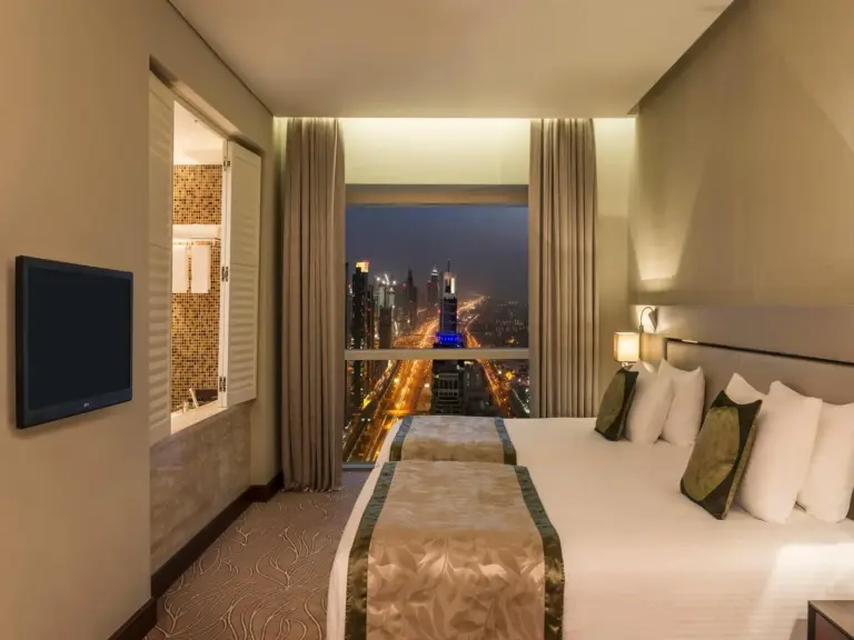 Modern bedroom at The Tower Plaza Hotel Dubai with a city skyline night view through large windows.