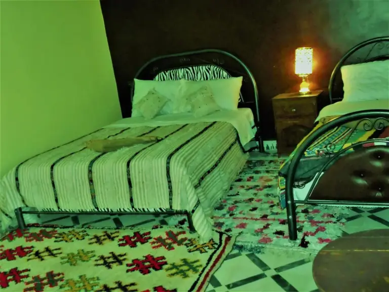 Habitación twin en Tifinagh Guest House en Merzouga con dos camas individuales, lámpara de noche y alfombras estampadas