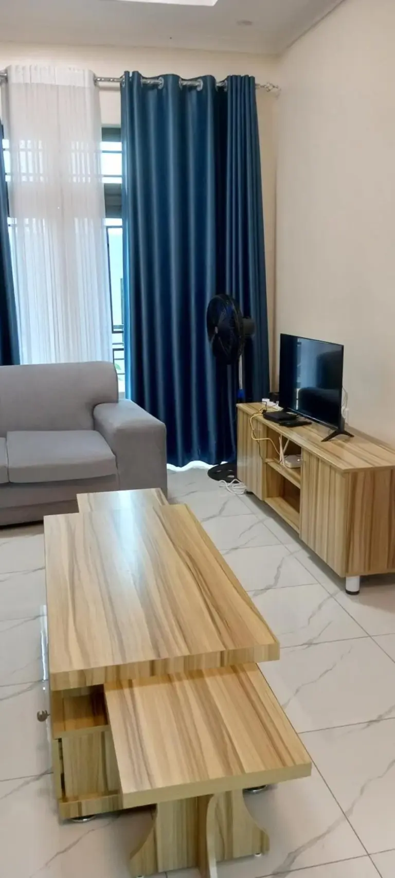 TV-Lounge im VENUS APARTMENT Kigali in Nyamata mit Vorhängen, einem Standventilator und einem kleinen Fernsehschrank.